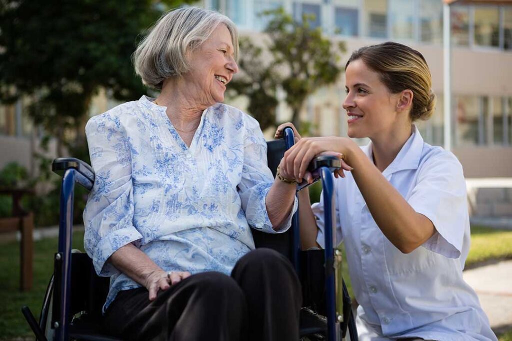 A care aide assists an older adult with a walker in a well-lit community hallway.